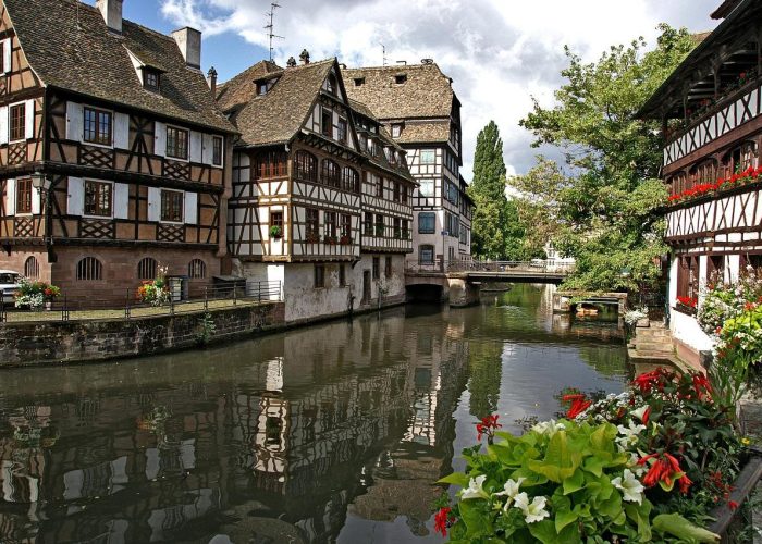 Picturesque scene of a canal with traditional half-timbered houses in Strasbourg, France, lined with flowers and trees, with reflections in the water and a small stone bridge in the background.