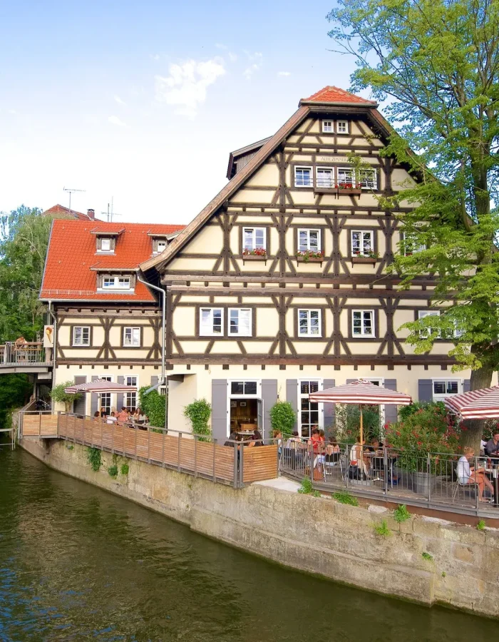 Sur une étroite bande de terre entourée d'eau se dresse un bâtiment traditionnel à colombages avec des places assises en plein air, où les gens mangent sous des parasols. Une église avec un haut clocher est visible à l'arrière-plan au milieu des arbres.