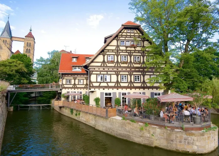 On a narrow strip of land surrounded by water stands a traditional half-timbered building with outdoor seating, where people eat under parasols. A church with a high steeple can be seen in the background among the trees.