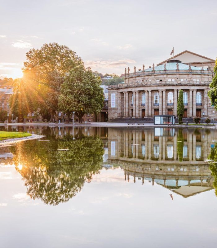 Le soleil se couche derrière un grand bâtiment néoclassique à colonnes, se reflétant dans un étang calme entouré d'arbres verts dans un parc.