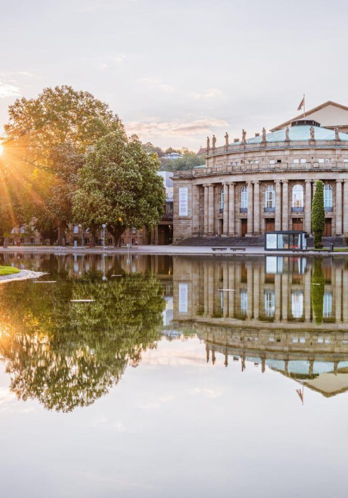 The sun sets behind a large neoclassical building with columns, reflected in a calm pond surrounded by green trees in a park.