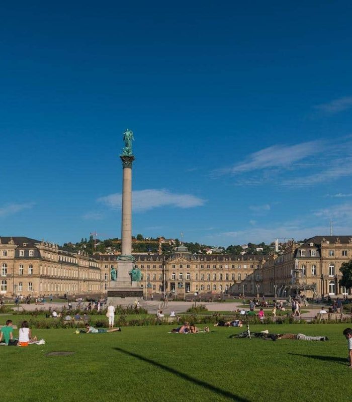 People relax on the green lawn of a town square, in front of a historic palace adorned with a tall column and a statue, under a clear blue sky.