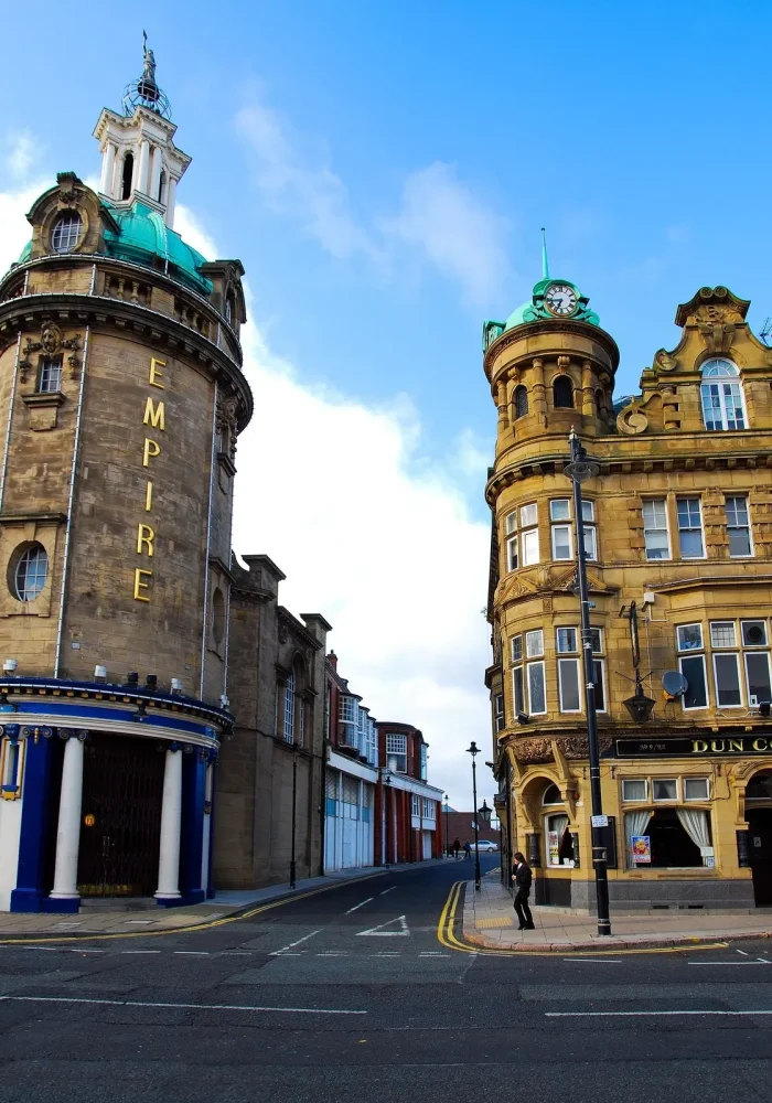 Deux bâtiments historiques ornés se dressent à l'angle d'une rue sous un ciel bleu ; celui de gauche est circulaire et porte l'inscription "EMPIRE", tandis que celui de droite porte l'inscription "DUN COW" et une personne marche à proximité.