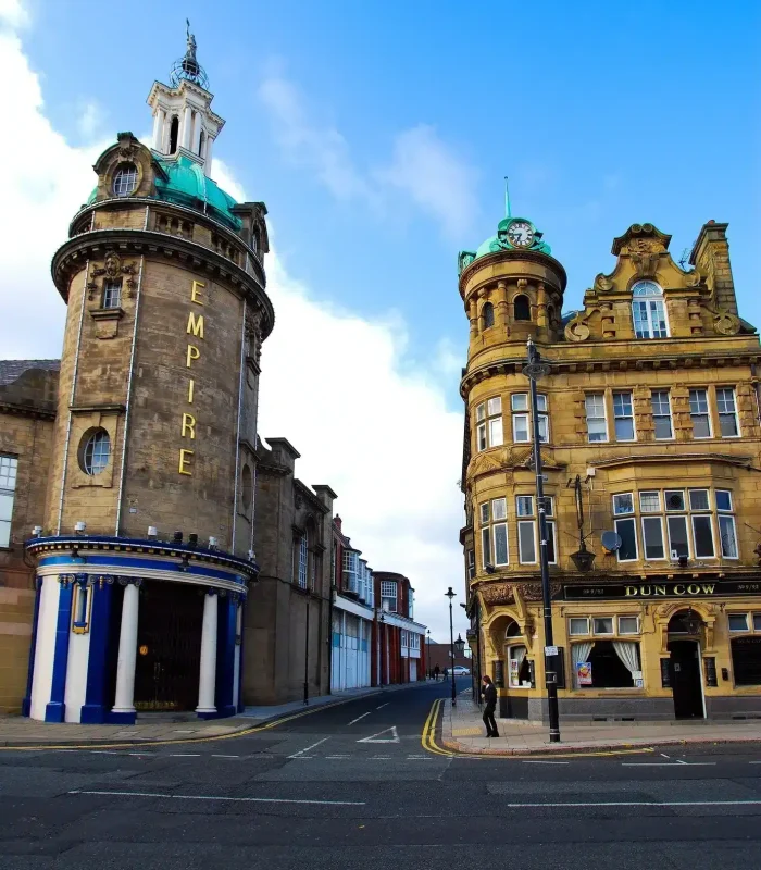 Two ornate historic buildings stand on a street corner under a blue sky; the one on the left is circular and bears the inscription "EMPIRE", while the one on the right bears the inscription "DUN COW" and a person is walking nearby.