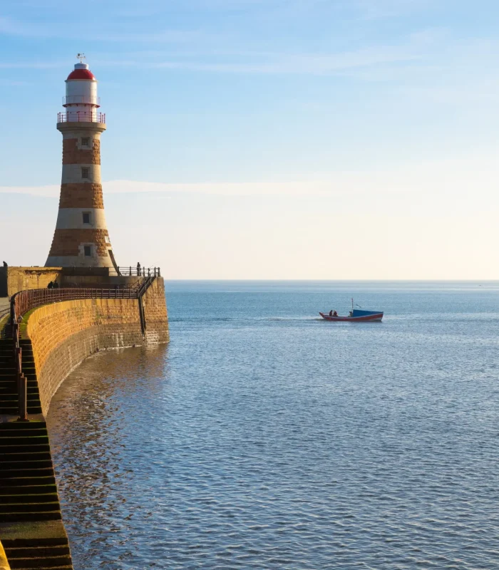 Un phare en pierre se dresse sur une jetée incurvée s'avançant dans une mer bleue et calme. Un petit bateau flotte près de la jetée sous un ciel clair, l'horizon étant visible au loin.