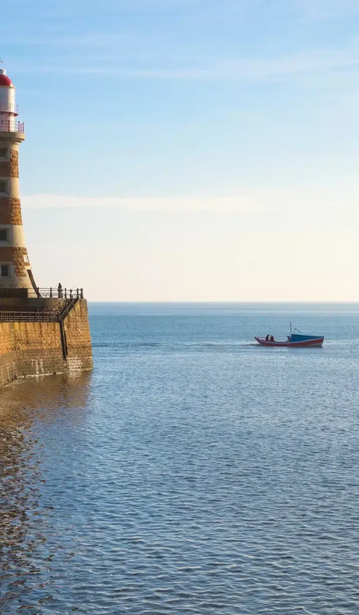A stone lighthouse stands on a curved jetty jutting out into a calm blue sea. A small boat floats near the jetty under a clear sky, with the horizon visible in the distance.