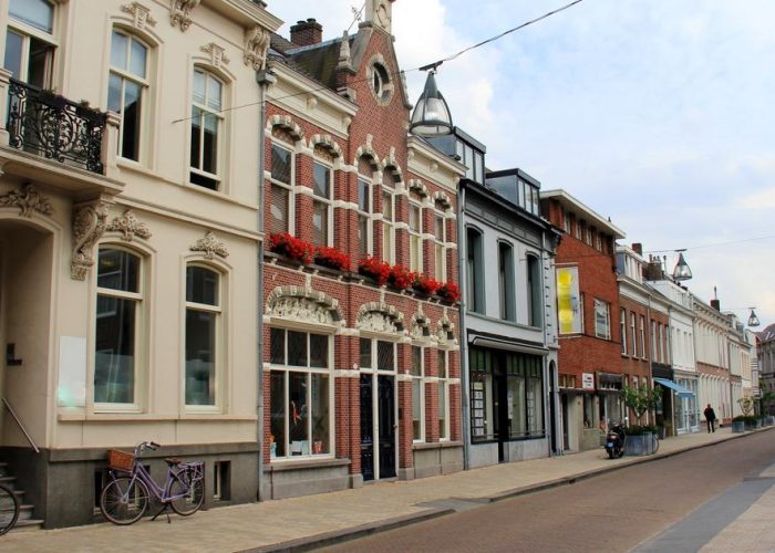 Une rue calme et étroite, bordée de bâtiments historiques européens ornés de grandes fenêtres, de balcons et de fleurs rouges. Des vélos sont garés le long du trottoir. Le ciel est partiellement nuageux.