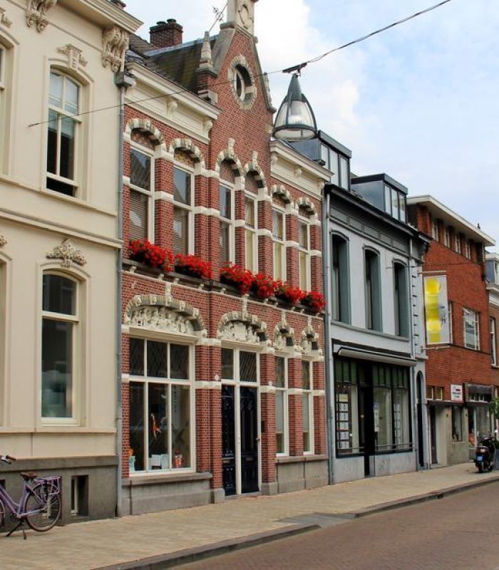 Une rue calme et étroite, bordée de bâtiments historiques européens ornés de grandes fenêtres, de balcons et de fleurs rouges. Des vélos sont garés le long du trottoir. Le ciel est partiellement nuageux.