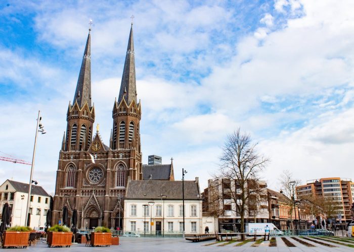 Une grande église de style gothique avec des flèches jumelles se dresse sur une vaste place urbaine, entourée d'arbres, de bâtiments modernes et d'un ciel bleu nuageux. Des personnes et des véhicules sont visibles sur la place ouverte.