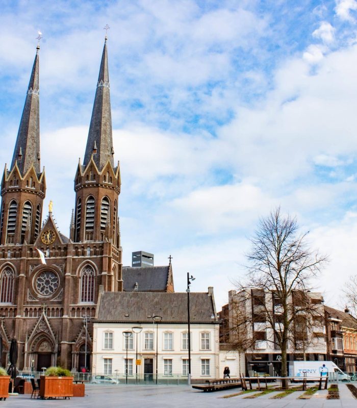 Une grande église de style gothique avec des flèches jumelles se dresse sur une vaste place urbaine, entourée d'arbres, de bâtiments modernes et d'un ciel bleu nuageux. Des personnes et des véhicules sont visibles sur la place ouverte.