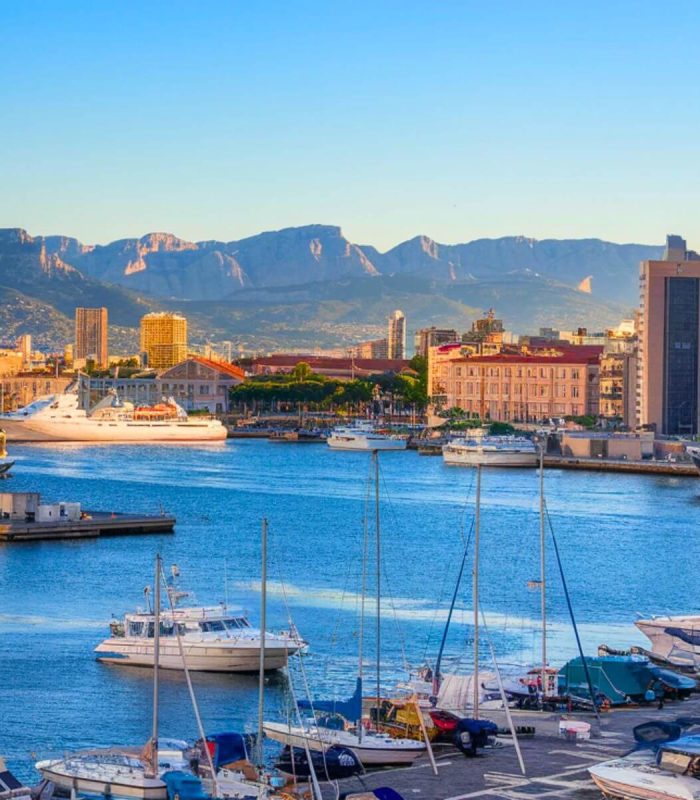 Bateaux et yachts amarrés dans un port de plaisance animé, avec des bâtiments urbains en arrière-plan, sur fond de montagnes dans un ciel bleu clair au coucher du soleil.