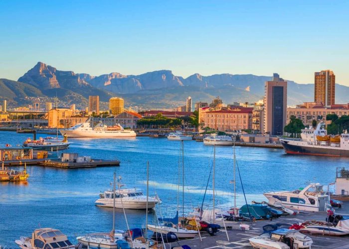 Boats and yachts moored in a bustling marina, with urban buildings in the background, against a backdrop of mountains in a clear blue sky at sunset.