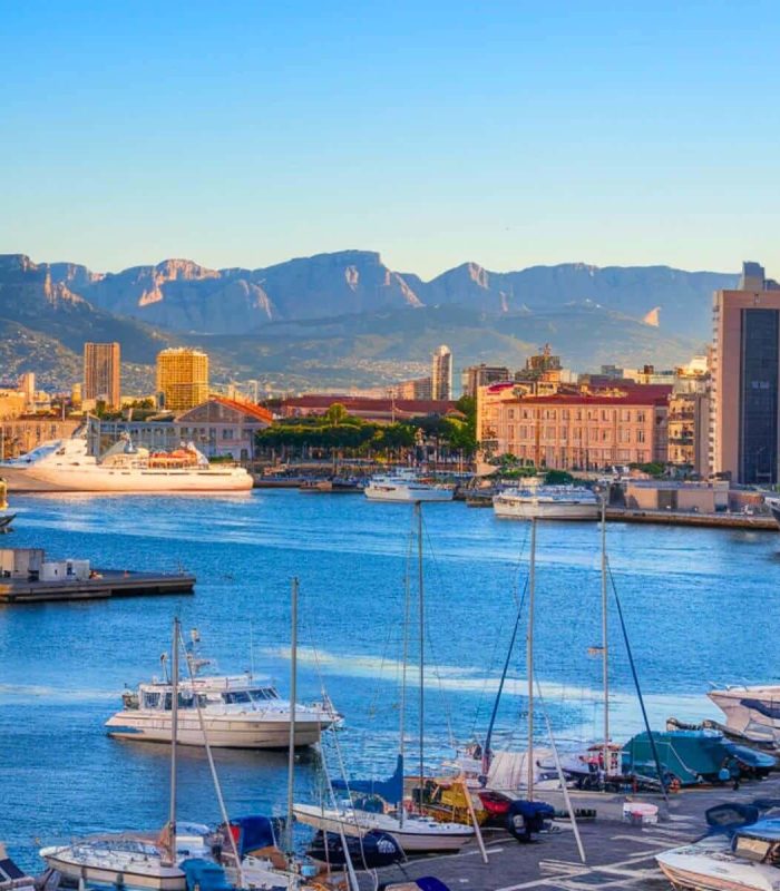 Bateaux et yachts amarrés dans un port de plaisance animé, avec des bâtiments urbains en arrière-plan, sur fond de montagnes dans un ciel bleu clair au coucher du soleil.