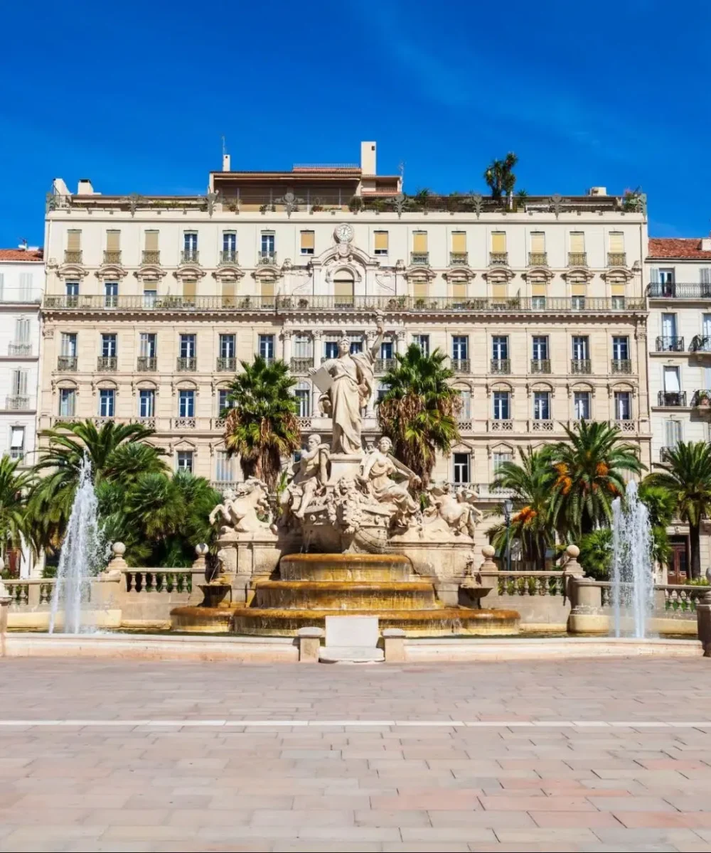 Une grande fontaine ornée de statues se dresse devant des palmiers et plusieurs bâtiments hauts et élégants, sous un ciel bleu clair, sur une vaste place de la ville.