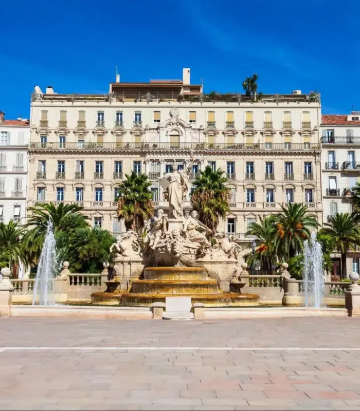 A large fountain adorned with statues stands in front of palm trees and several tall, elegant buildings, under a clear blue sky, in a vast town square.