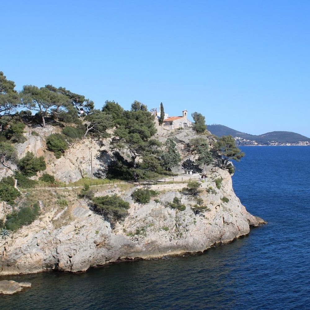 Falaise côtière rocheuse couverte d'arbres et de végétation, surplombant une mer bleue et calme. Un bâtiment au toit rouge et quelques cyprès se trouvent au sommet de la falaise. Des collines lointaines sont visibles sous un ciel bleu clair.