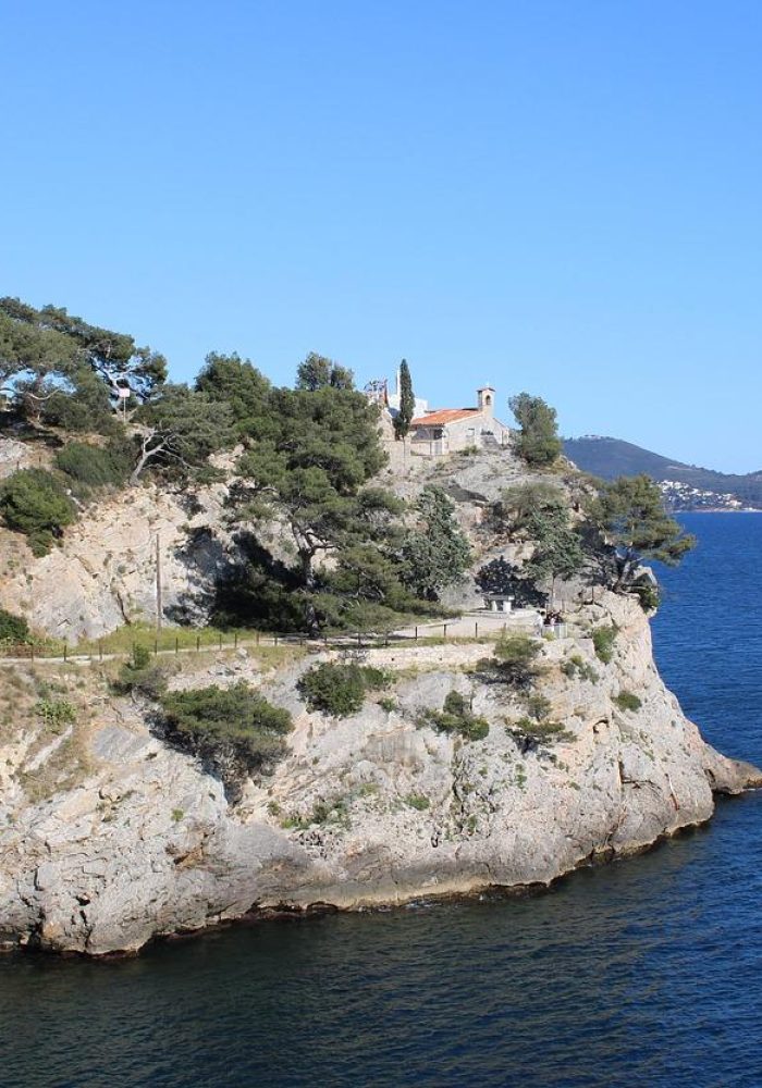 A rocky coastal cliff covered in trees and vegetation, overlooking a calm blue sea. A red-roofed building and a few cypress trees stand at the top of the cliff. Distant hills are visible under a clear blue sky.