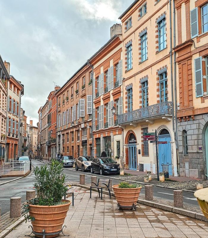 A picturesque European street lined with tall, colourful buildings with shutters and balconies. Potted plants and benches adorn the pavement, while a few people walk by and cars are parked along the pavement.