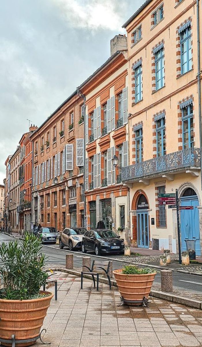 Une rue européenne pittoresque bordée de grands immeubles colorés avec des volets et des balcons. Des plantes en pot et des bancs ornent le trottoir, tandis que quelques personnes marchent et que des voitures sont garées le long du trottoir.