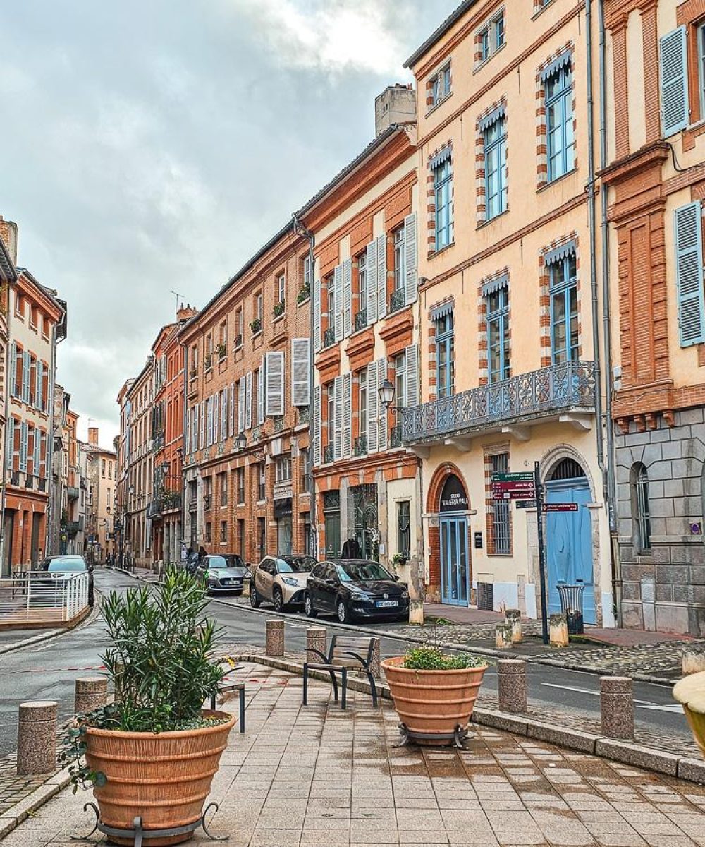Une rue européenne pittoresque bordée de grands immeubles colorés avec des volets et des balcons. Des plantes en pot et des bancs ornent le trottoir, tandis que quelques personnes marchent et que des voitures sont garées le long du trottoir.