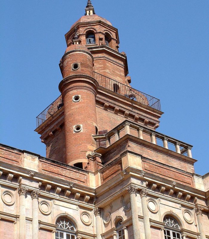 Vue rapprochée d'une tour historique en briques avec des sections cylindriques et rectangulaires, des fenêtres arquées et des détails architecturaux décoratifs, sur fond de ciel bleu clair.
