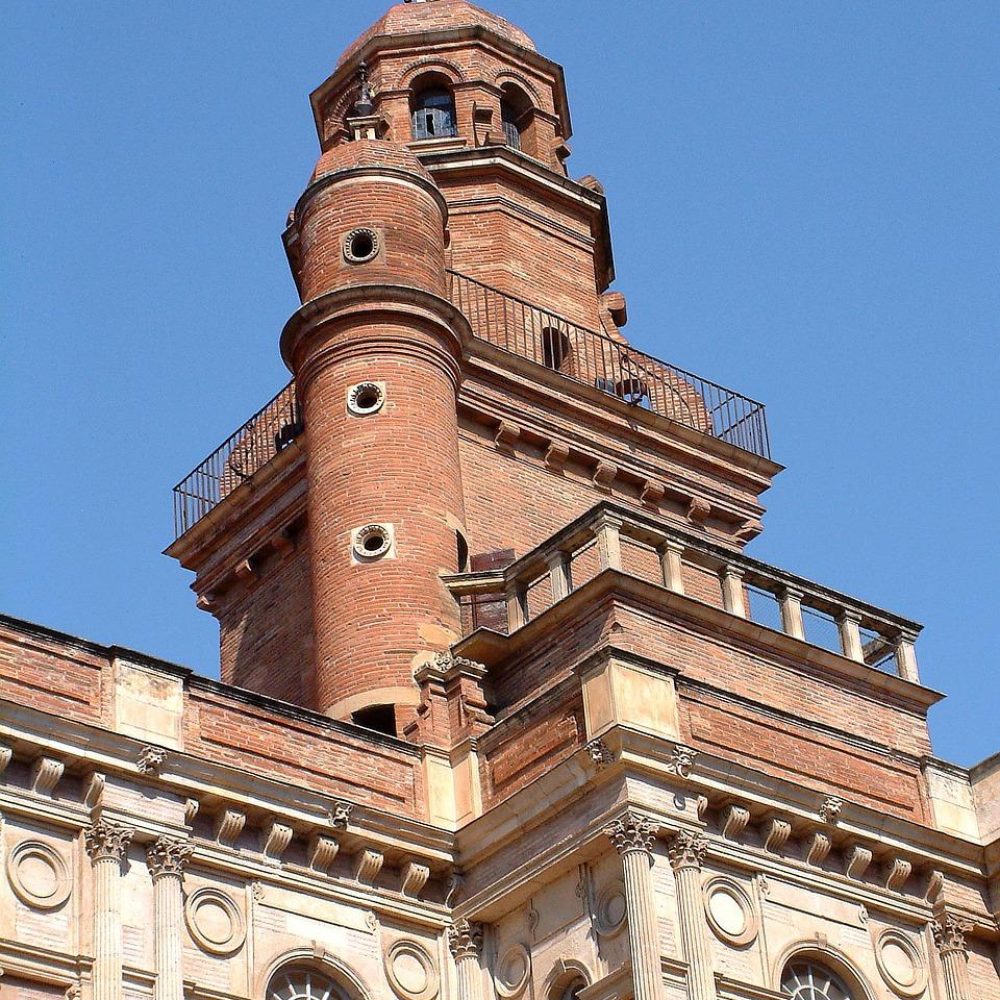 Vue rapprochée d'une tour historique en briques avec des sections cylindriques et rectangulaires, des fenêtres arquées et des détails architecturaux décoratifs, sur fond de ciel bleu clair.