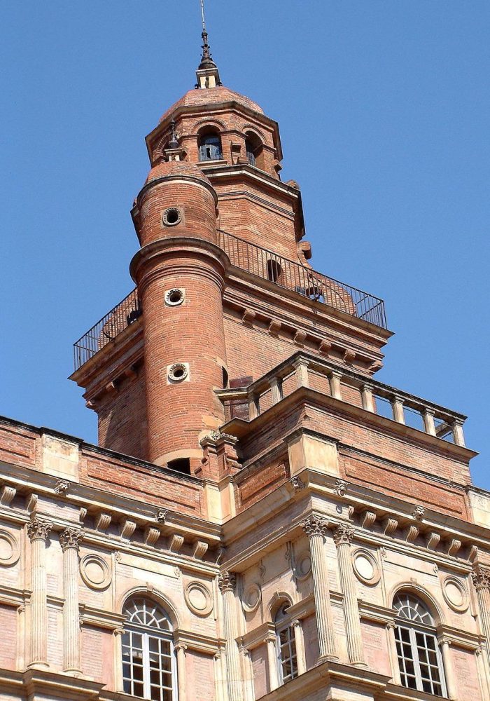 Close-up view of a historic brick tower with cylindrical and rectangular sections, arched windows and decorative architectural details, against a backdrop of light blue sky.