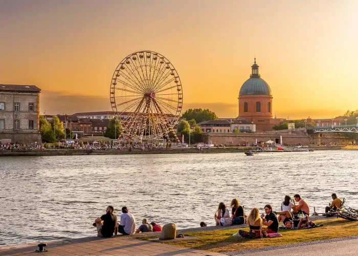 People are sitting on the grassy bank enjoying the sunset, with a Ferris wheel, historic dome and city buildings in the background on the other side of the water. The sky is golden and festive.