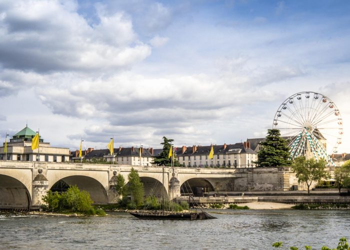 Un pont de pierre enjambe une rivière avec des bâtiments et des arbres en arrière-plan ; une grande roue s'élève au-dessus des toits sous un ciel partiellement nuageux. Des drapeaux jaunes ornent le pont.