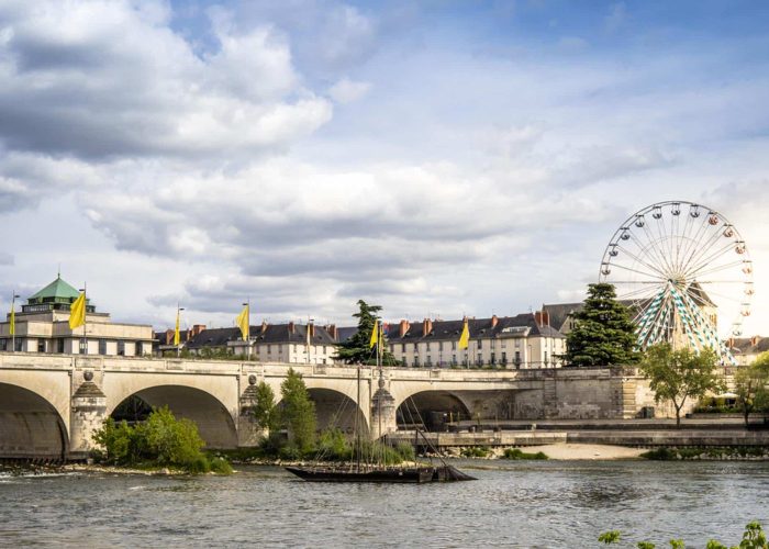 A stone bridge spans a river with buildings and trees in the background; a Ferris wheel rises above the rooftops under a partly cloudy sky. Yellow flags adorn the bridge.