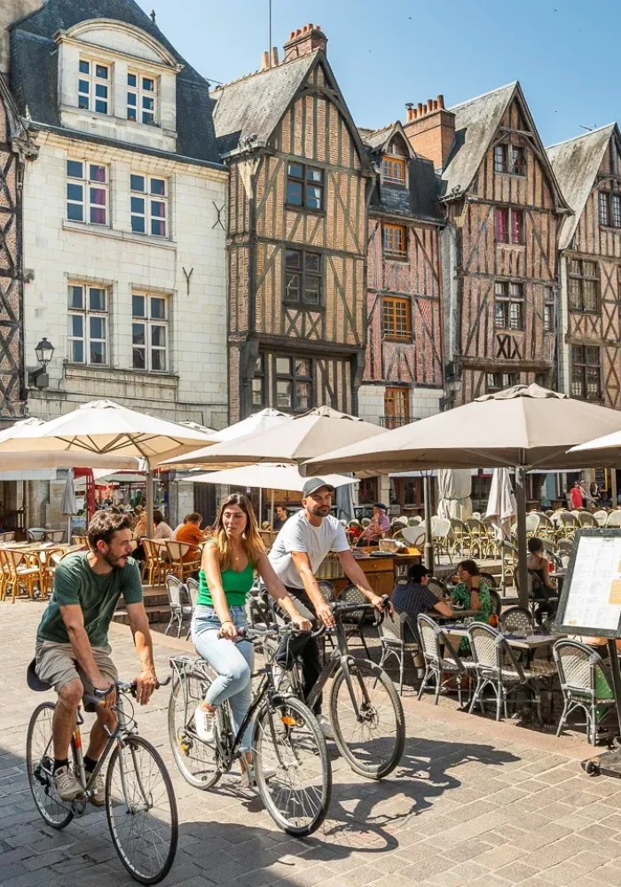 Une place européenne animée avec des gens qui mangent dans des cafés en plein air sous des parasols blancs, des cyclistes qui passent et des bâtiments historiques à colombages en arrière-plan par une journée ensoleillée.