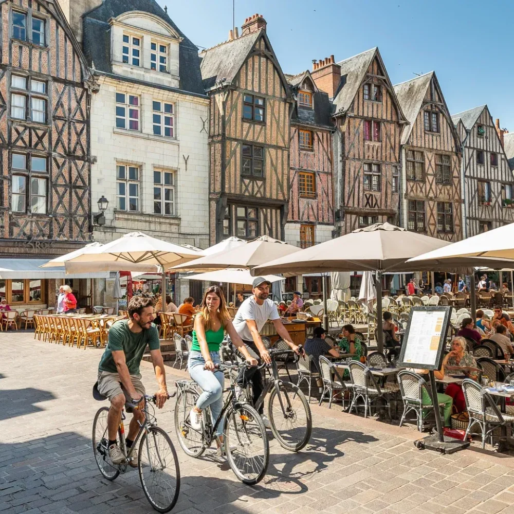 Une place européenne animée avec des gens qui mangent dans des cafés en plein air sous des parasols blancs, des cyclistes qui passent et des bâtiments historiques à colombages en arrière-plan par une journée ensoleillée.