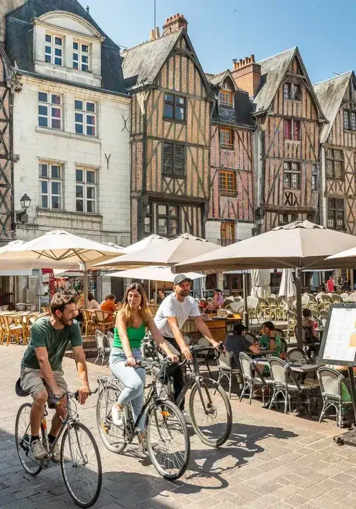 Une place européenne animée avec des gens qui mangent dans des cafés en plein air sous des parasols blancs, des cyclistes qui passent et des bâtiments historiques à colombages en arrière-plan par une journée ensoleillée.