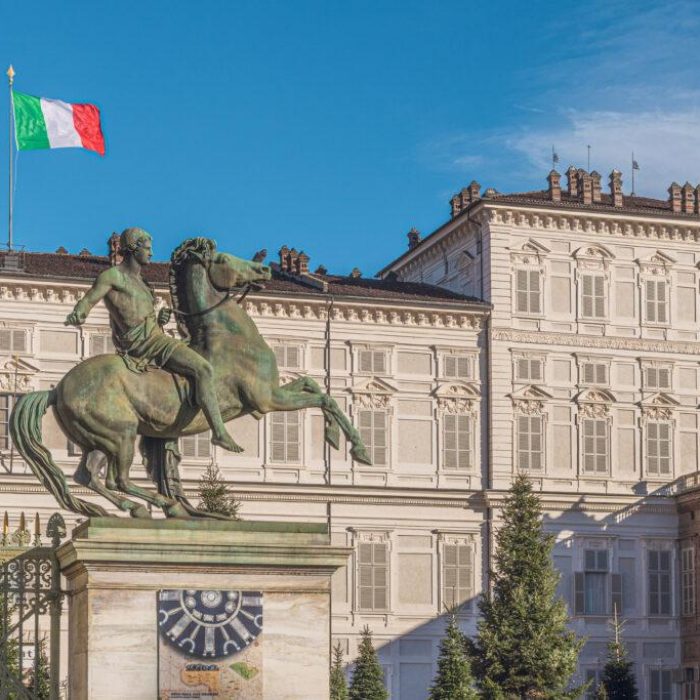 Une statue équestre en bronze se dresse devant un grand bâtiment historique doté de nombreuses fenêtres. Un drapeau italien flotte sur le toit dans un ciel bleu clair.