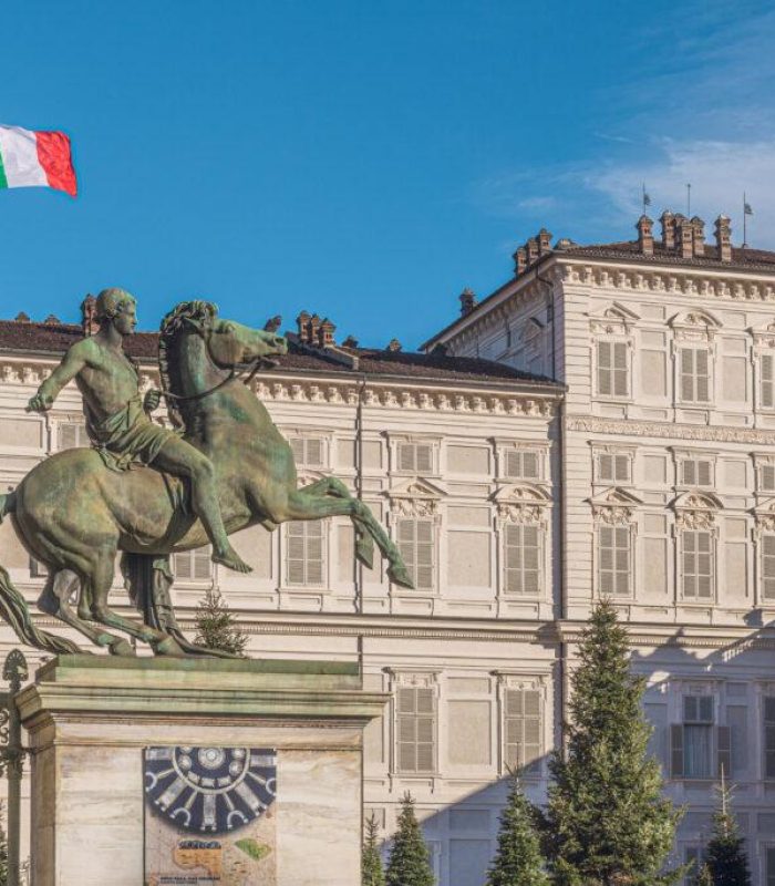 Une statue équestre en bronze se dresse devant un grand bâtiment historique doté de nombreuses fenêtres. Un drapeau italien flotte sur le toit dans un ciel bleu clair.