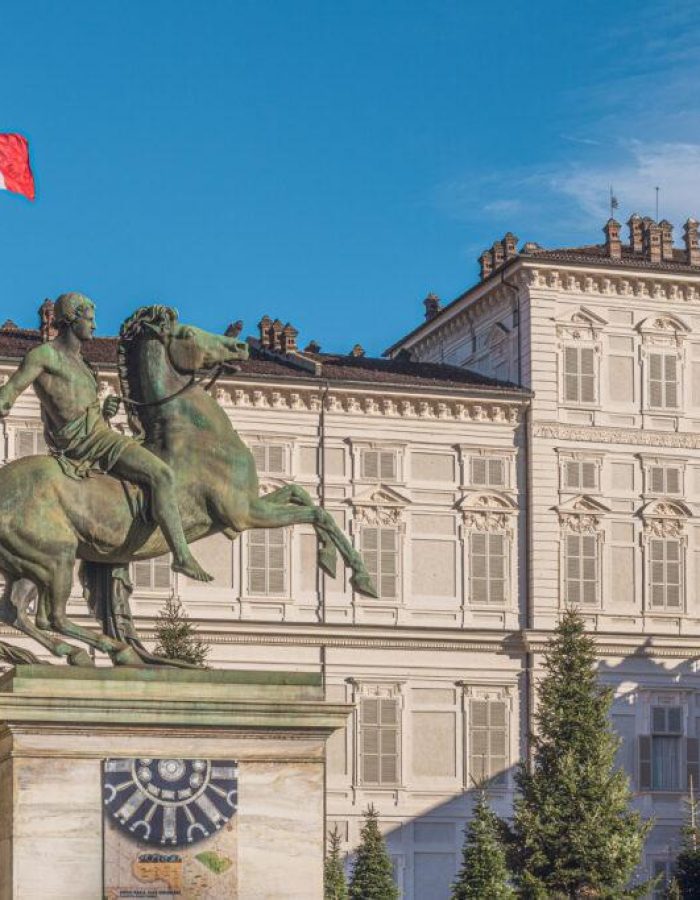Une statue équestre en bronze se dresse devant un grand bâtiment historique doté de nombreuses fenêtres. Un drapeau italien flotte sur le toit dans un ciel bleu clair.