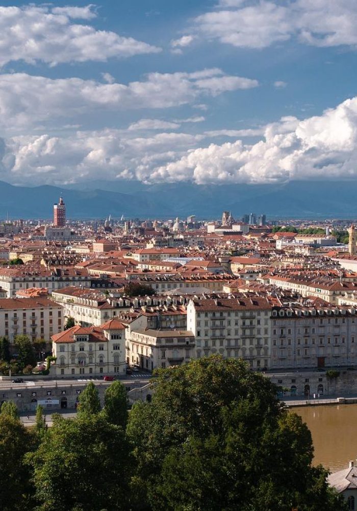 Vue panoramique de Turin, Italie, avec des bâtiments historiques aux toits rouges, la Mole Antonelliana et des montagnes en arrière-plan, sous un ciel partiellement nuageux. Une rivière longe la ville, bordée d'arbres.
