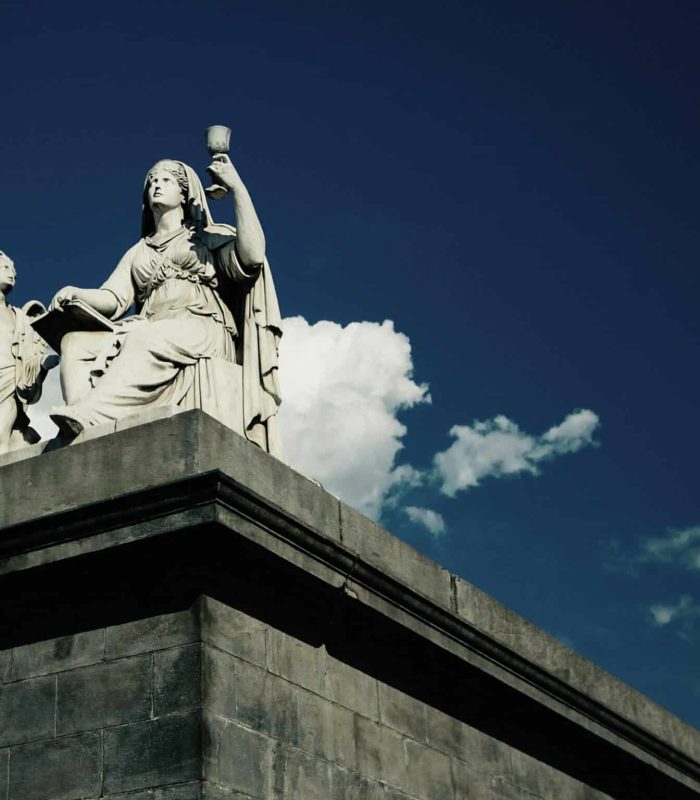 Une statue en pierre blanche représentant une femme assise tenant une coupe et un enfant debout est posée sur un grand piédestal en pierre, dans un ciel d'un bleu profond parsemé de nuages. Une partie d'un bâtiment est visible sur la droite.