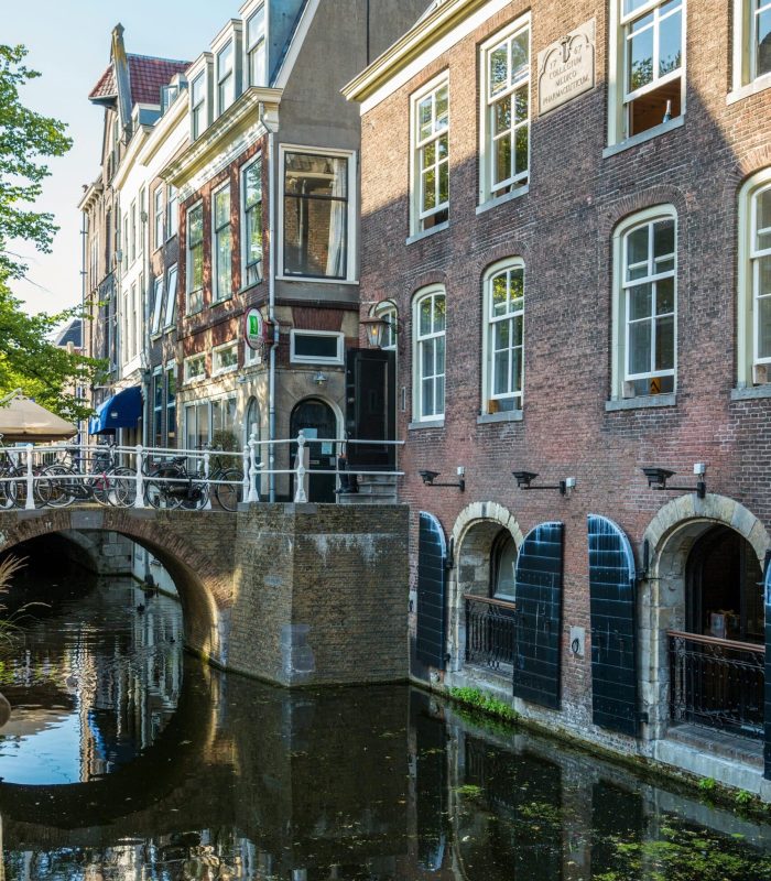 Un canal étroit longe des bâtiments en briques avec des portes cintrées au niveau de l'eau. Un petit pont avec des rambardes traverse le canal, et la scène est éclairée par la lumière du soleil avec des reflets dans l'eau.