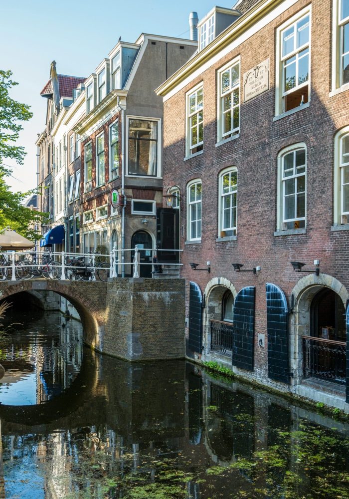 Un canal étroit longe des bâtiments en briques avec des portes cintrées au niveau de l'eau. Un petit pont avec des rambardes traverse le canal, et la scène est éclairée par la lumière du soleil avec des reflets dans l'eau.