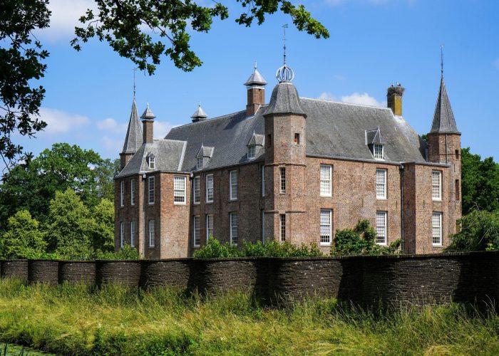 Un grand château historique en briques, aux multiples tourelles et aux toits pentus, se dresse derrière un mur de pierre, entouré d'herbe verte et d'arbres, sous un ciel bleu parsemé de nuages.
