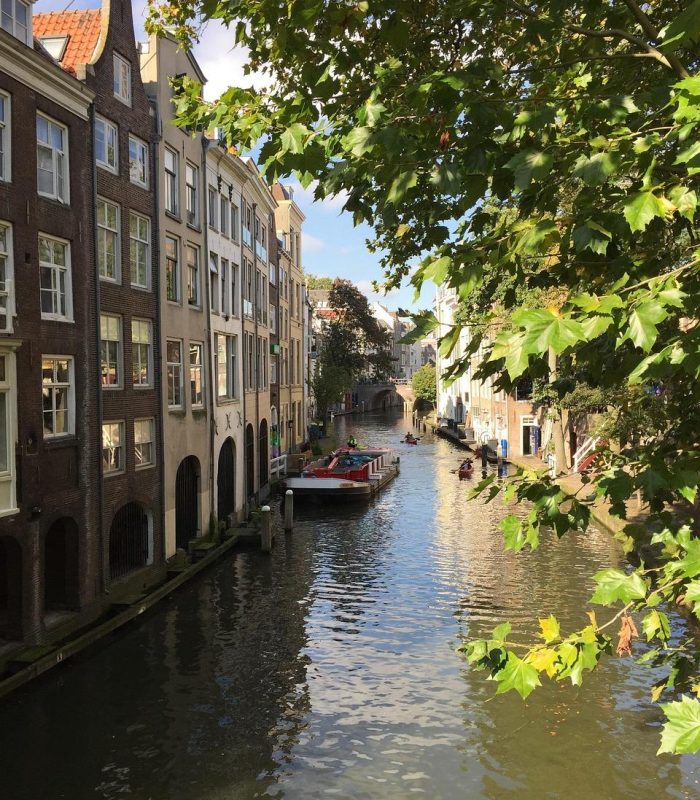 Un canal étroit bordé de hauts bâtiments historiques des deux côtés, un bateau amarré au centre, et des arbres verts feuillus qui se dressent au-dessus de l'eau, sous un ciel lumineux et partiellement nuageux.