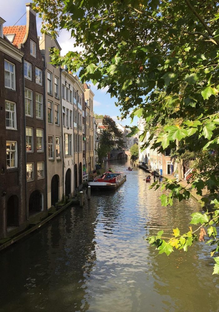 Un canal étroit bordé de hauts bâtiments historiques des deux côtés, un bateau amarré au centre, et des arbres verts feuillus qui se dressent au-dessus de l'eau, sous un ciel lumineux et partiellement nuageux.