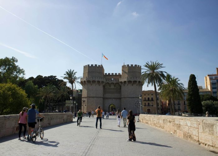 Des personnes traversent à pied ou à vélo un pont de pierre en direction de la porte médiévale Torres de Serranos, une porte de ville à deux tours, à Valence, en Espagne, avec des palmiers et un ciel d'un bleu éclatant à l'arrière-plan.