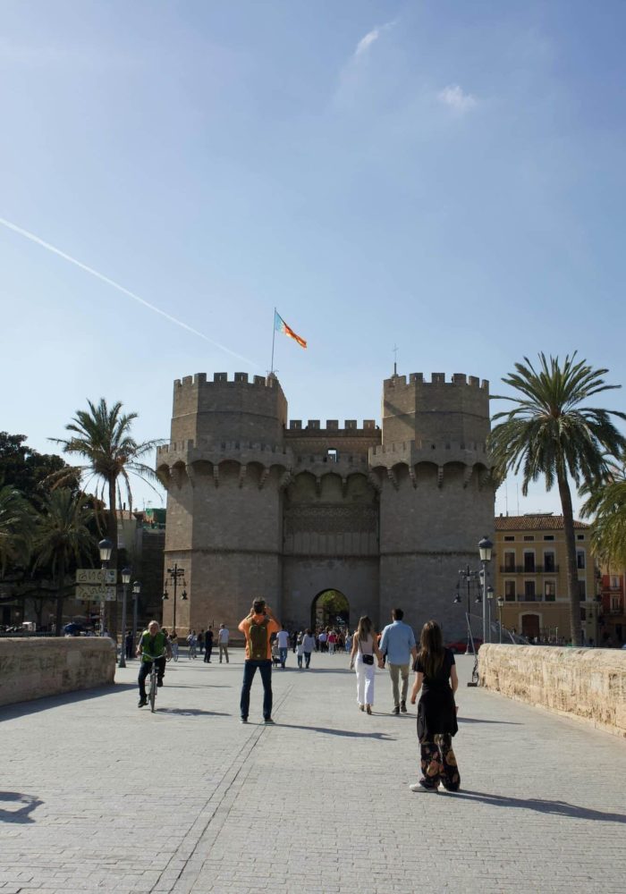 Des personnes traversent à pied ou à vélo un pont de pierre en direction de la porte médiévale Torres de Serranos, une porte de ville à deux tours, à Valence, en Espagne, avec des palmiers et un ciel d'un bleu éclatant à l'arrière-plan.