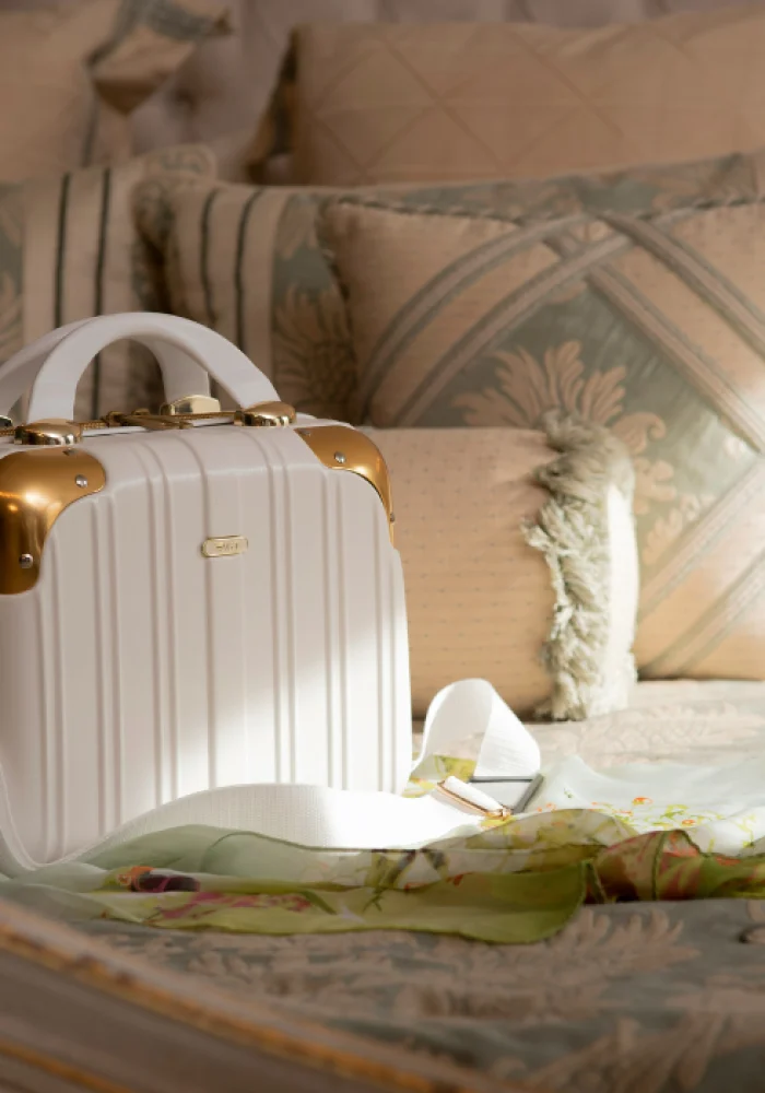 Une petite valise de voyage blanche avec des accents dorés et une sangle blanche est posée sur un lit orné d'oreillers à motifs et d'un foulard floral vert clair, parfait pour ceux qui cherchent à stocker des bagages sécurisés pendant leur voyage.