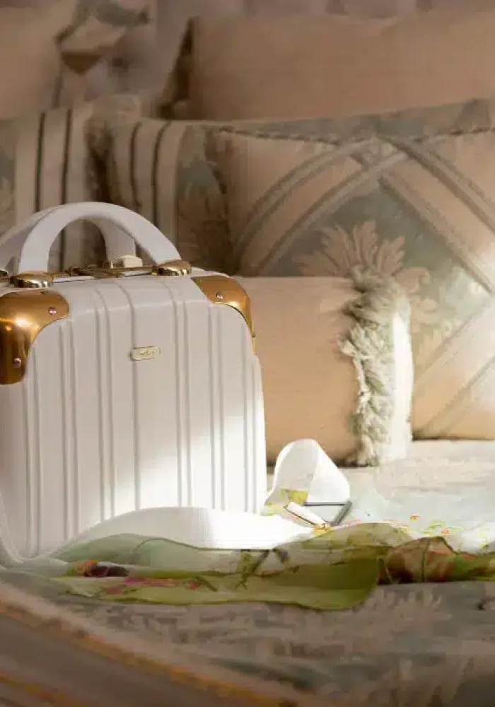 A small white travel suitcase with gold accents and a white strap sits on a bed adorned with patterned pillows and a light green floral scarf, perfect for those looking to store secure luggage while travelling.