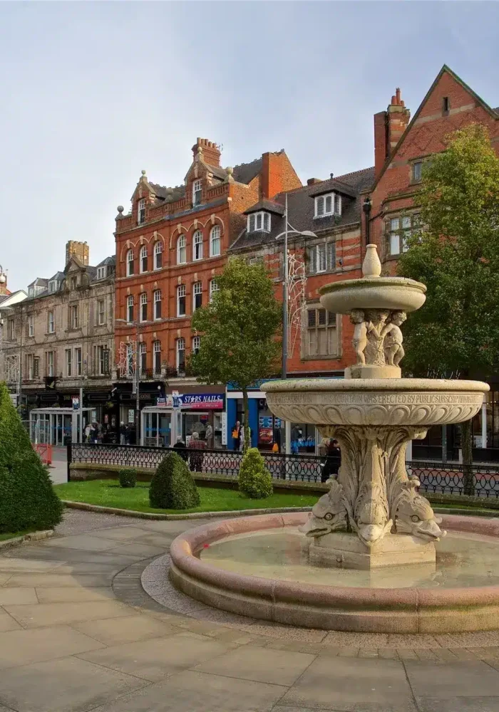 Une fontaine en pierre avec des figures sculptées se trouve sur une place pavée, entourée de buissons taillés, avec des bâtiments historiques en brique et des magasins en arrière-plan sous un ciel clair.