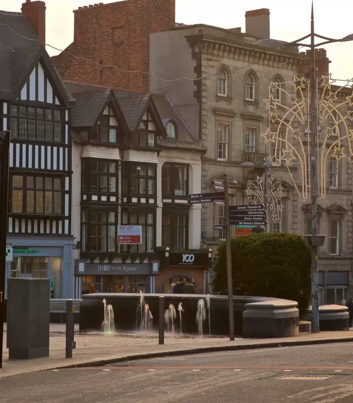 Scène de rue au coucher du soleil avec des bâtiments historiques, dont des façades à colombages et en pierre, des boutiques, une petite fontaine, des lampadaires et quelques personnes marchant sur le trottoir.