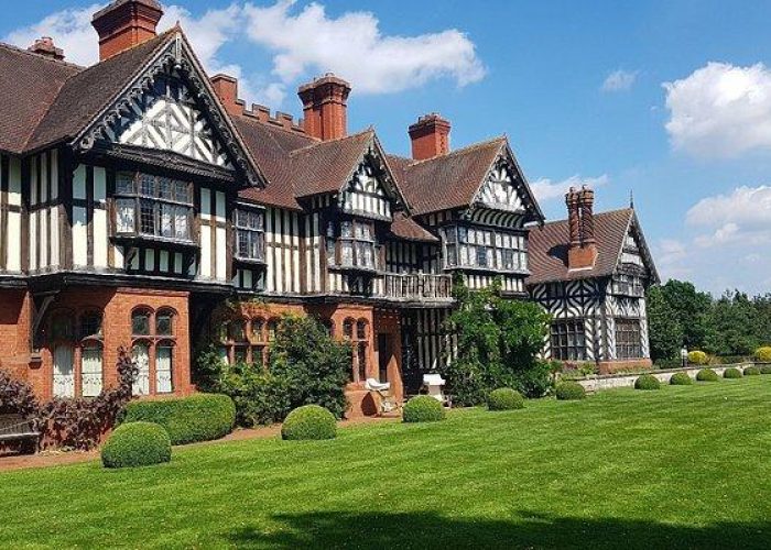 Un grand manoir historique de style Tudor avec une charpente en bois, de hautes cheminées et des pelouses vertes entretenues sous un ciel bleu brillant avec des nuages épars.